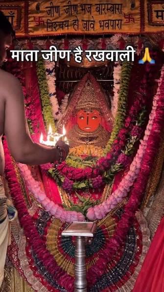A priest offers the aarti lamp before Jeen Mata, decorated with red and pink garlands; the screen text reads 'Mata Jeen hai rakhwali'
