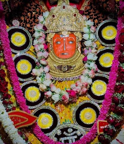 Close darshan of Jeen Bhawani's sindoor-orange face with silver eyes, golden crown and rose and jasmine garlands, framed by decorated silver shields