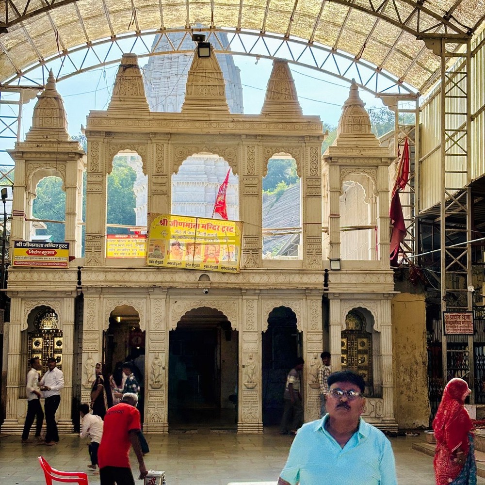 The carved cream-stone entrance gateway of the Shri Jeen Mata Mandir Trust temple, with domed pavilions, a saffron flag, a yellow trust banner and devotees in the courtyard under a curved canopy