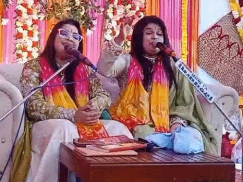 Two women bhajan singers in yellow and red bandhej dupattas sing devotional songs into microphones on a flower-decorated stage, one raising her hand